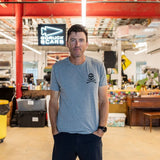 A man wearing the GORUCK Tribe Tee - Tri-Blend stands in a vibrant industrial workspace with a “GORUCK SCARS” sign, embodying the authentic community spirit of the GORUCK Tribe.