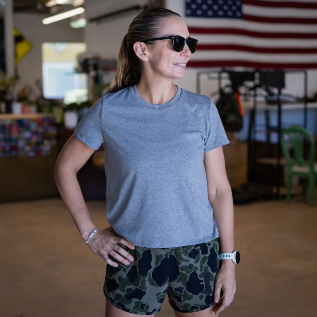 A woman wearing a grey Tri-BlendX t-shirt and 18″ length camouflage shorts stands in a workshop with an American flag hanging in the background.