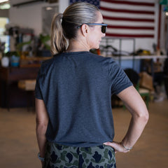 A woman seen from behind wearing an 18″ length Tri-BlendX dark gray t-shirt and camouflage pants, with an American flag hanging in the background.