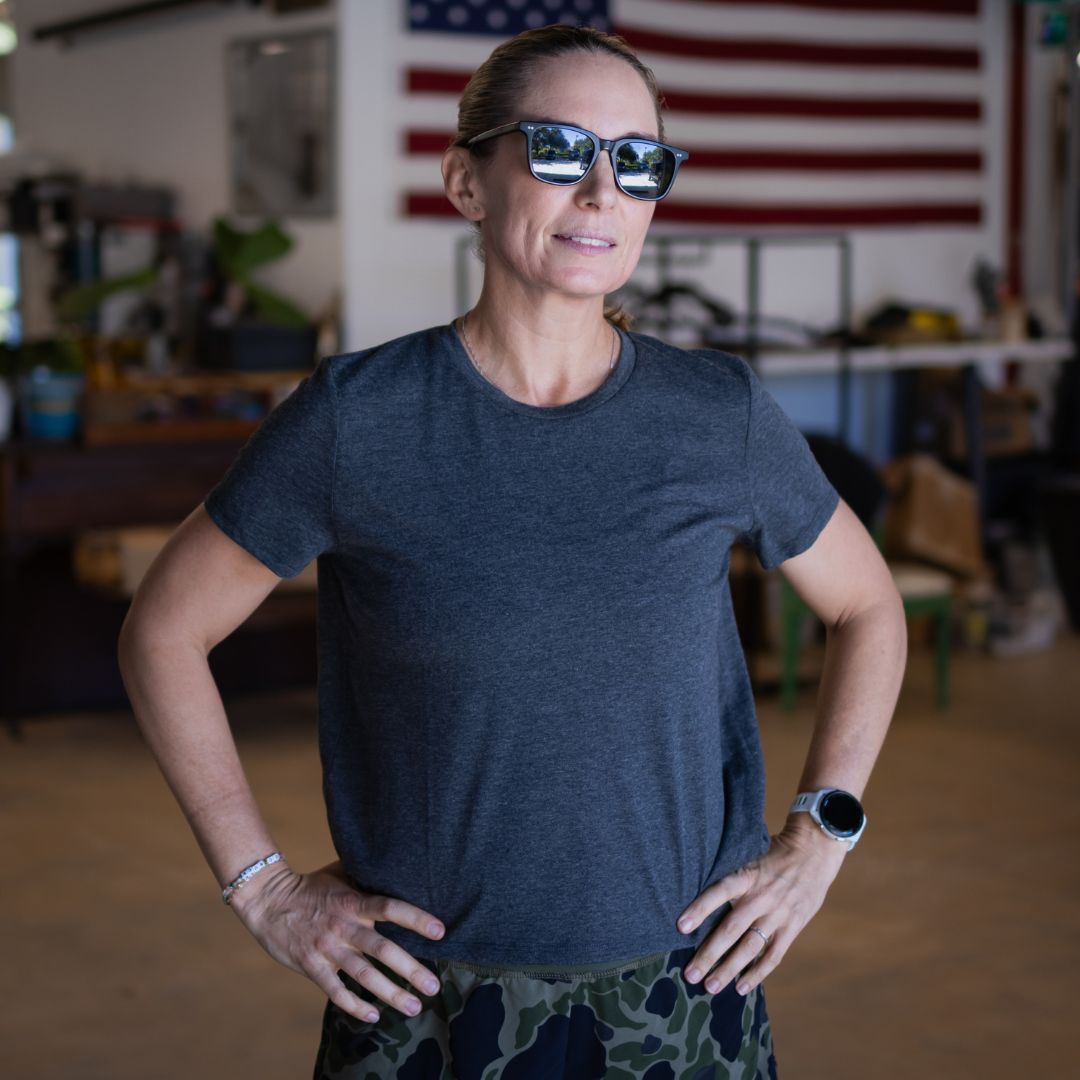 A woman wearing sunglasses and a charcoal Tri-BlendX t-shirt with an 18″ length stands with her hands on her hips in front of an American flag.