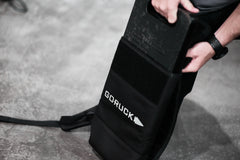 Someone inserts a 30LB plate into the RPC Long backpack on a gray floor, showcasing the ergonomic lumbar support of the RPC Long + 30LB (Long) Plate Bundle for added comfort during intense workouts.