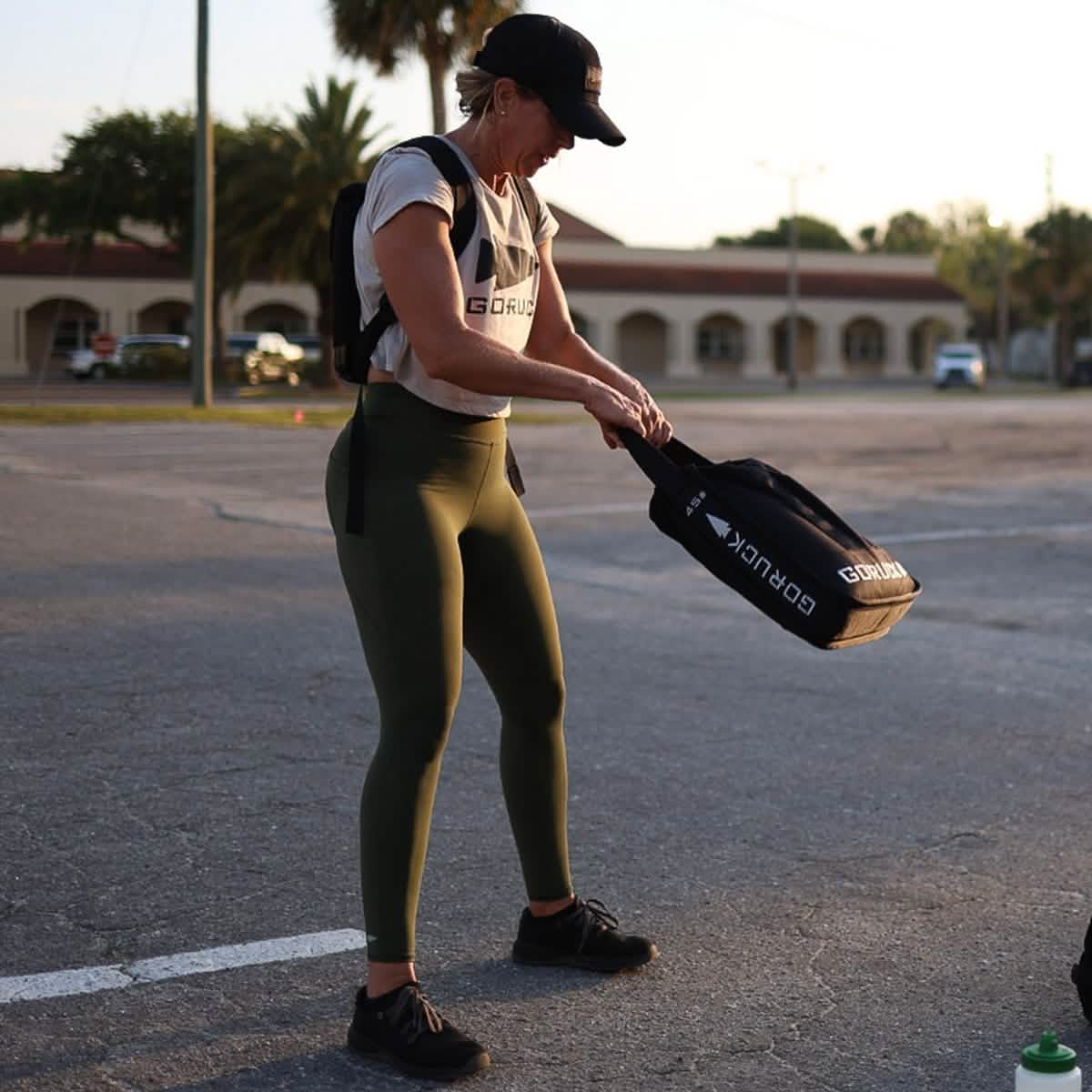 A woman wearing a cap, workout shirt, and green leggings adjusts a paddle in a pickleball paddle cover outdoors. She stands in a sunlit parking lot with trees and buildings in the background, highlighting her core strength, with a GORUCK Sand Jerry Can sports water bottle on the ground nearby.