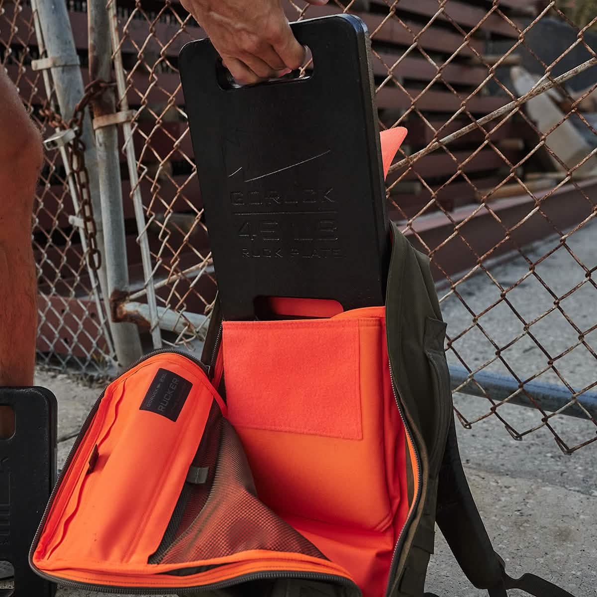 A person carefully places a GORUCK Ruck Plates weighing 45 LB into an open backpack featuring an orange interior, crafted for toughness and durability. The backpack, called the Rucker, rests on the ground near a chain-link fence.