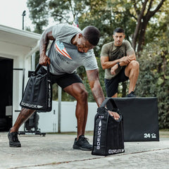 Two men are enhancing their core strength outdoors. One is doing squats with a weighted Sand Jerry Can from GORUCK, made from Special Forces Grade materials, while the other crouches on a platform in the driveway. Nearby, trees and bushes provide a natural backdrop to their intense workout.