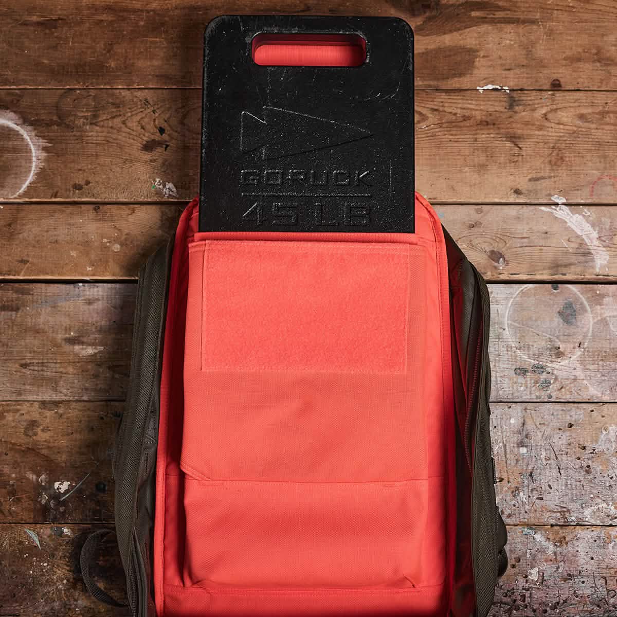A 45 LB GORUCK Ruck Plate, meticulously crafted in the USA, comes with a carrying handle and is partially tucked into an open backpack with a vivid red interior, placed on a wooden floor.