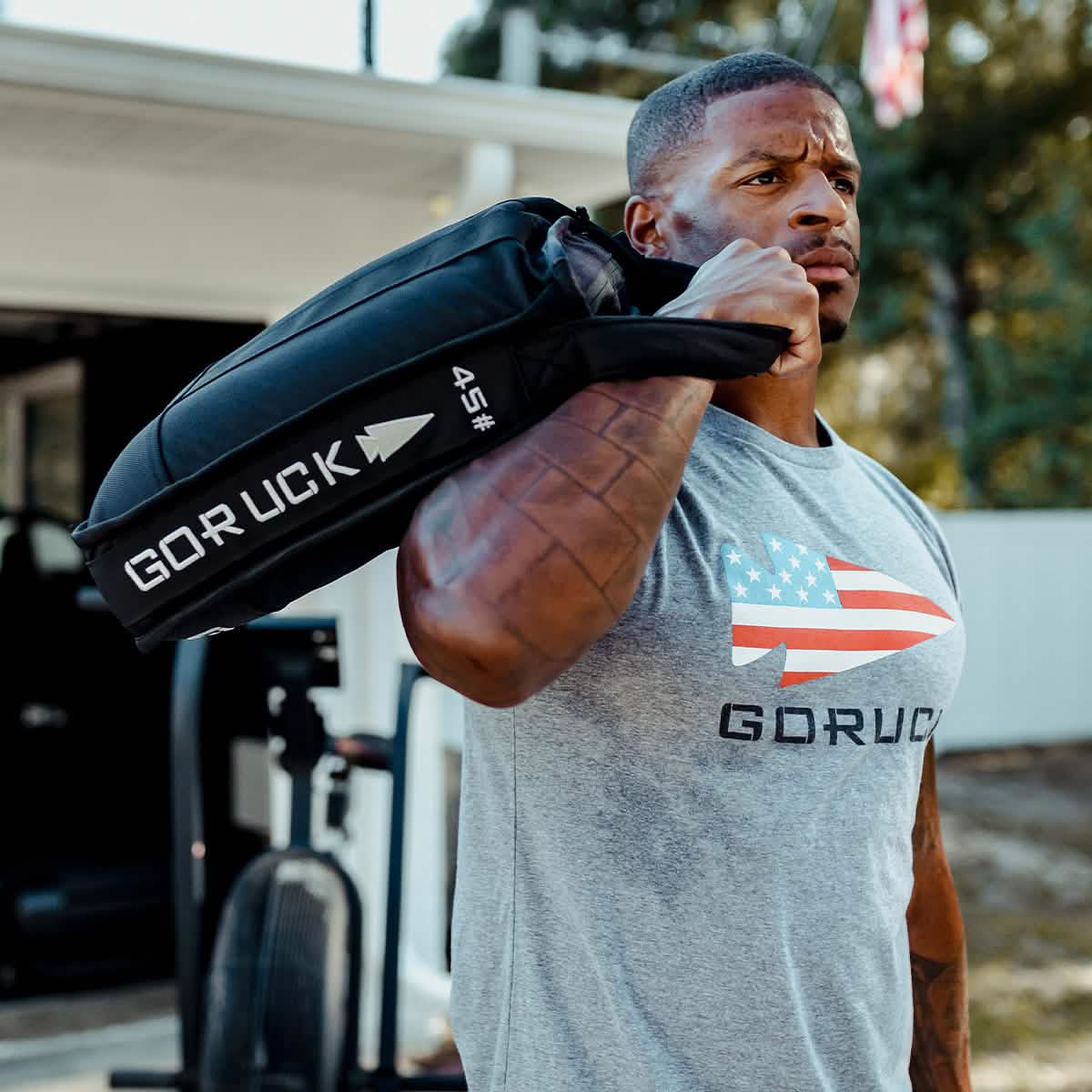 A man wearing a gray GORUCK shirt with an American flag design carries a black GORUCK Sand Jerry Can, crafted from Special Forces Grade materials, on his shoulder. He stands near outdoor exercise equipment, focused and determined to build core strength.