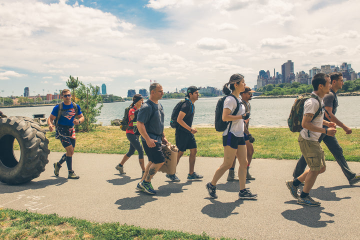 A group of people with backpacks walk on a riverside path in a city park on a sunny day.