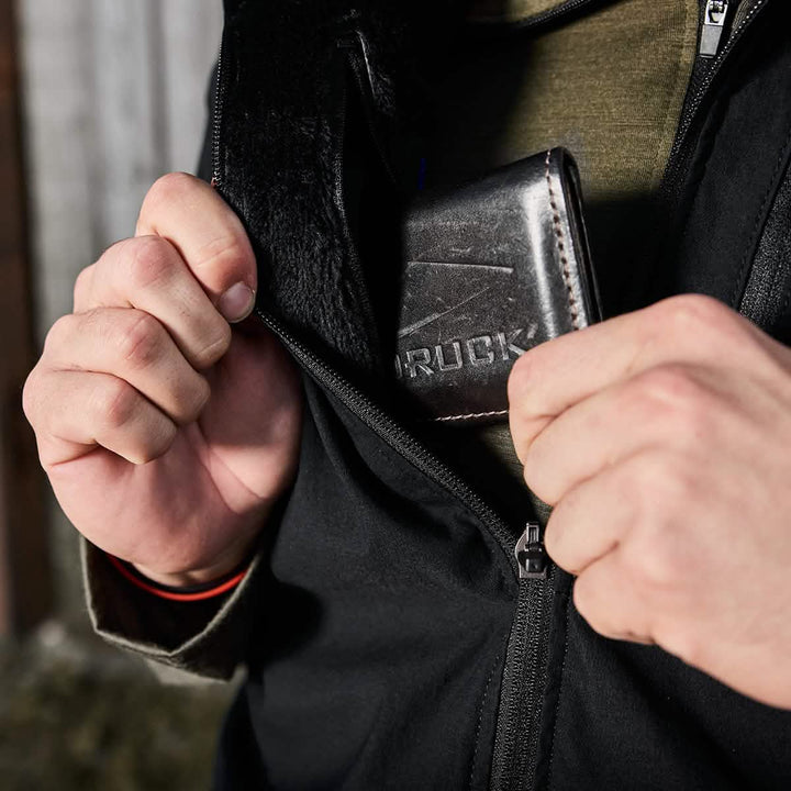 A person zips up the Vest of Power - ToughDry® + Fleece, a sleek black piece from GORUCK, while holding a dark leather wallet with the brand's name partially visible. The green shirt underneath hints at an environmentally conscious choice, blending style with subtle eco-awareness.
