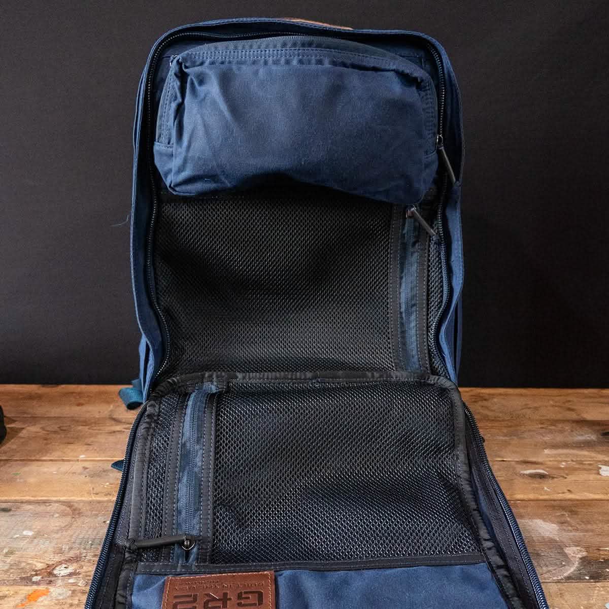 An open blue backpack rests on a wooden surface, highlighting its interior compartments. Featuring mesh pockets for organization and a zippered pouch near the top, this GR2 Heritage backpack by GORUCK with its Alligator Leather Front Panel stands out. The dark background enhances the allure of its sleek design.
