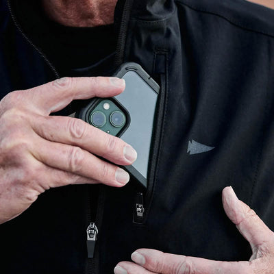 A person wearing the GORUCK Vest of Power in black, crafted from ToughDry® and Fleece, carefully slides a smartphone with a dual camera into the zippered chest pocket. The environmentally conscious design highlights a small triangular logo, drawing attention to their hands and the phone.
