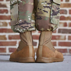 A close-up of a person wearing camouflage pants and lightweight rucking boots captures the scene. The GORUCK MACV-1 Hi-Speed - High Top military boots, AR670-1 compliant, are paired perfectly with the tucked-in pants. The person stands before a brick wall, drawing focus to their lower legs and feet.