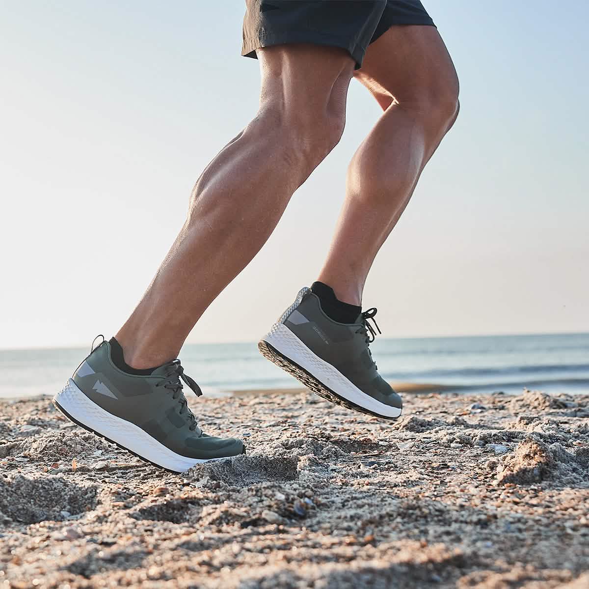 A person running on a sandy beach in GORUCK's Men's Rough Runner - Earth shoes, with the ocean and a clear sky as their backdrop.