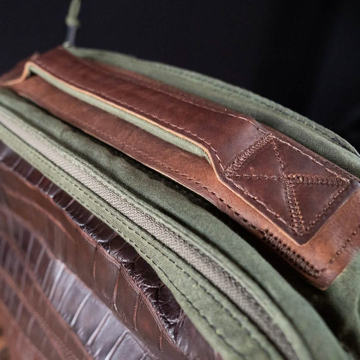 Detailed view of a GORUCK GR1 Heritage - Alligator Leather Front Panel bag handle crafted from brown leather and green canvas. The handle showcases intricate stitching, with leather and canvas materials prominently displayed, evoking a rustic and durable appearance that harks back to vintage canvas backpacks.