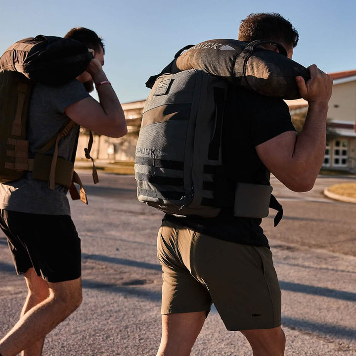 Two individuals, sporting GORUCK backpacks equipped with padded hip belts and carrying bags over their shoulders, walk along a paved surface. Clad in athletic attire, they proceed smoothly despite their heavy loads, while a large building stands visible in the background under a clear sky.