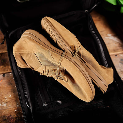 A pair of tan MACV-1 Hi-Speed - Mid Top sneakers by GORUCK, featuring a lightweight and rugged design for versatile casual or outdoor wear, sits neatly inside an open black suitcase on a wooden surface.