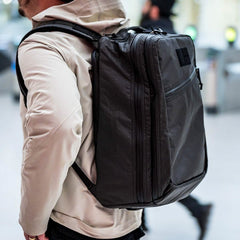 A person wearing a beige jacket carries the GR2 USA x Carryology - Ultra Black backpack, crafted from UHMWPE fabric, in an indoor setting.