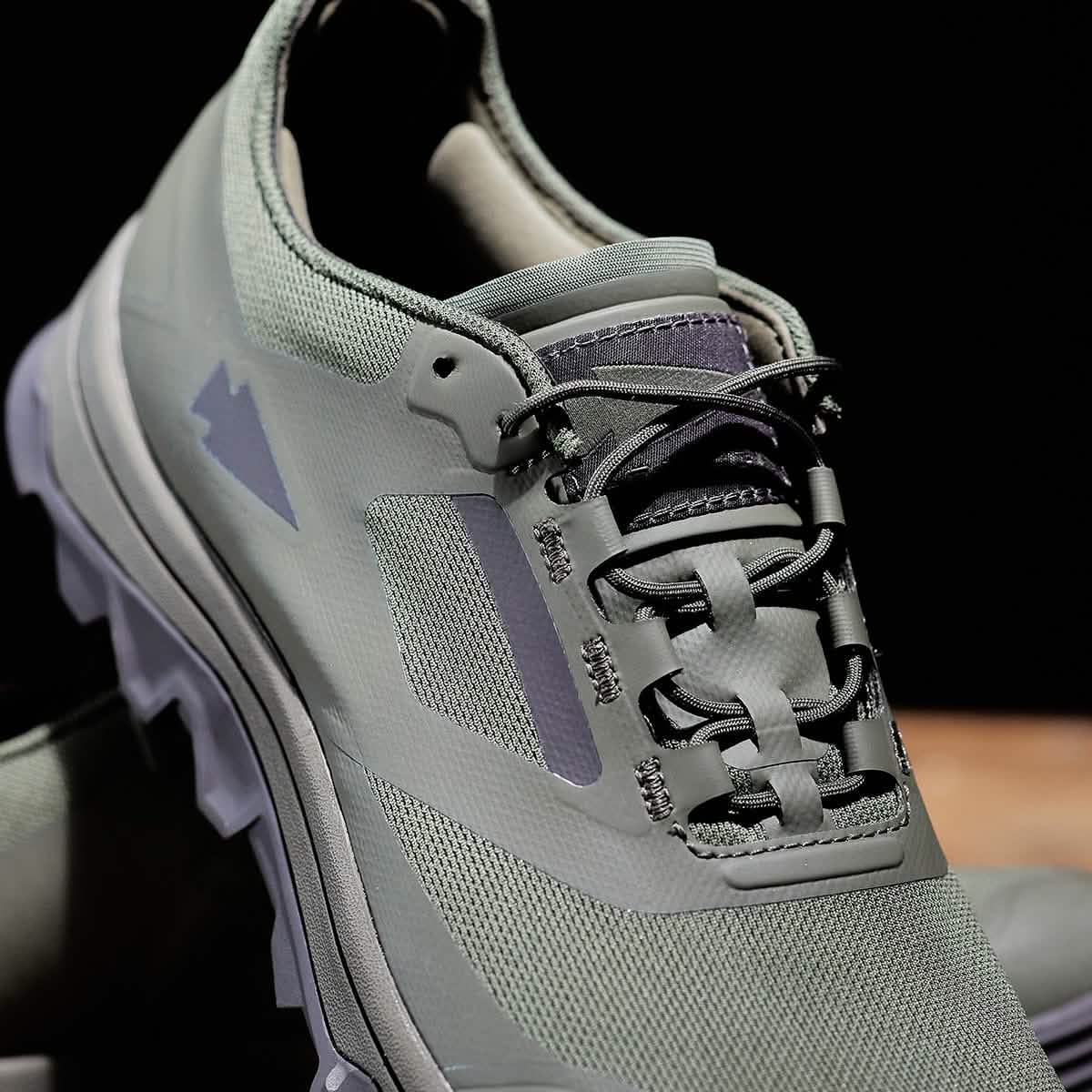 A close-up of the GORUCK Mackall in Earth + Forged Iron showcases a trail shoe with a textured surface and intricate lacing. The shoe bears a mountain-shaped logo and is equipped with a rugged triple compound outsole, perfect for outdoor activities. The dark background accentuates the shoe's features.
