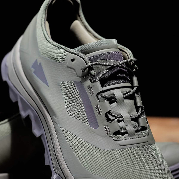 A close-up of the GORUCK Mackall in Earth + Forged Iron showcases a trail shoe with a textured surface and intricate lacing. The shoe bears a mountain-shaped logo and is equipped with a rugged triple compound outsole, perfect for outdoor activities. The dark background accentuates the shoe's features.