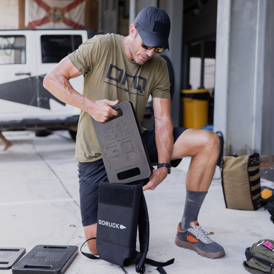 A man in a green t-shirt and baseball cap kneels on the ground, inserting a rectangular weight plate into a Long Ruck Plate Carrier.