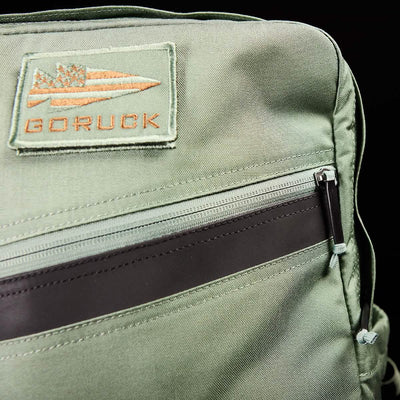 A detailed view of the green KR1 kids rucksack from GORUCK showcases a branded patch near the top. This backpack includes padded straps for comfort and features a front horizontal zipper accentuated by a dark brown strip, all against a black backdrop. It's ideal for stylishly carrying school supplies.