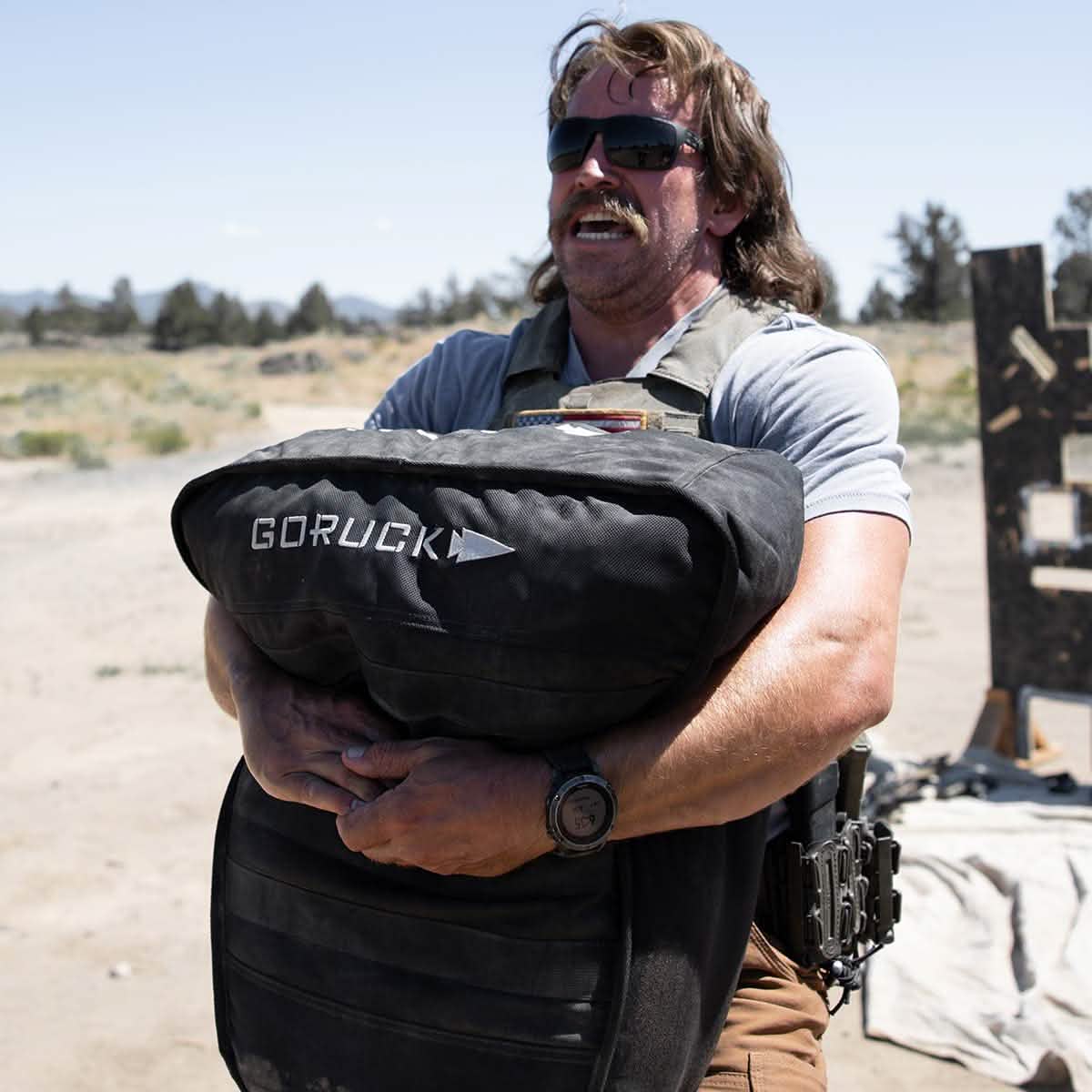 A person wearing sunglasses and a mustache is outdoors, sporting a tactical vest and holding a large Sand Tombstones bag from GORUCK, featuring a multilayer closure system. The setting implies rugged, physically demanding activities such as loaded carries under clear skies.