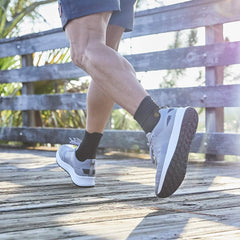 A person strolls along the wooden boardwalk, wearing GORUCK's Men's Rough Runner sneakers in Speed Grey + White with black socks, as sunlight filters through the railings—a true Rough Runner moment.