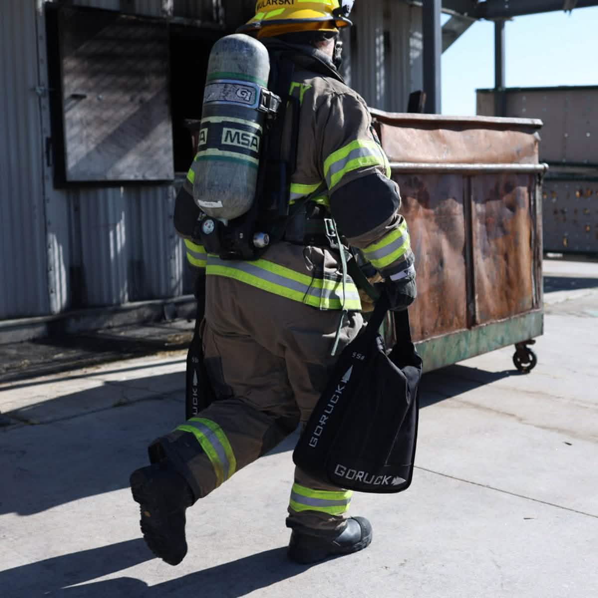 A firefighter in full gear, including a helmet and oxygen tank, showcases core strength by walking while carrying two GORUCK Sand Jerry Cans near a large metal dumpster. The reflective stripes on the uniform, crafted from Special Forces Grade materials, are clearly visible. The scene seems to be located near a training facility or fire station.