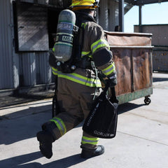 A firefighter in full gear, including a helmet and oxygen tank, showcases core strength by walking while carrying two GORUCK Sand Jerry Cans near a large metal dumpster. The reflective stripes on the uniform, crafted from Special Forces Grade materials, are clearly visible. The scene seems to be located near a training facility or fire station.