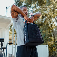 A person in a gray T-shirt and black shorts exercises outdoors, lifting a Sand Jerry Can from GORUCK, crafted with Special Forces Grade materials to enhance core strength. An American flag and trees are visible in the background, creating a patriotic workout setting.