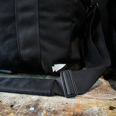 A close-up of a black GORUCK Kit Bag, complete with a sturdy shoulder strap, rests on a rustic wooden surface. The bag features a small, white embroidered arrow logo and includes an adjustable metal buckle on the strap, making it perfect for travel adventures.