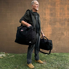 A man stands on grass in front of a brick wall, holding two large GORUCK Kit Bags with shoulder straps. He wears a dark jacket, gray pants, and brown boots as he gazes into the distance.