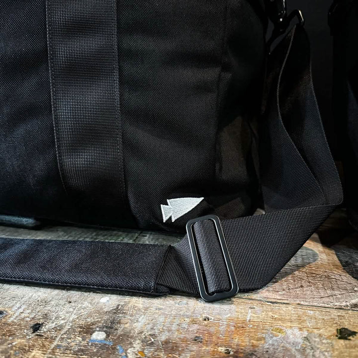A close-up of a black GORUCK Kit Bag, complete with a sturdy shoulder strap, rests on a rustic wooden surface. The bag features a small, white embroidered arrow logo and includes an adjustable metal buckle on the strap, making it perfect for travel adventures.