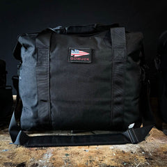 A black GORUCK Kit Bag, complete with an American flag patch on the front, rests with the stoic demeanor of a Special Forces Flight Bag on a wooden surface against a dark background.