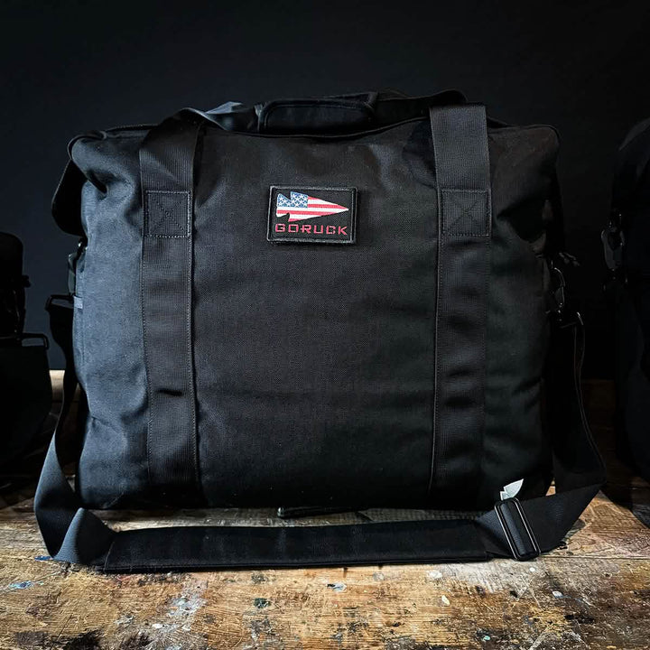 A black GORUCK Kit Bag, complete with an American flag patch on the front, rests with the stoic demeanor of a Special Forces Flight Bag on a wooden surface against a dark background.