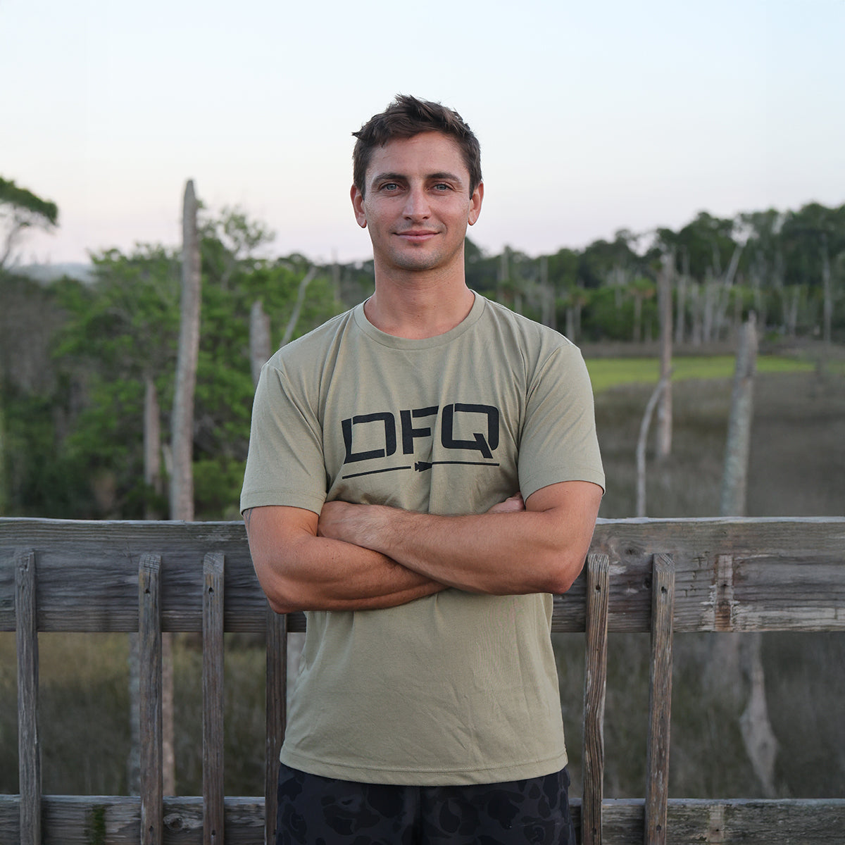 A man stands outside on a wooden deck, arms crossed, wearing the DFQ Tee - Tri-Blend, which features the bold 