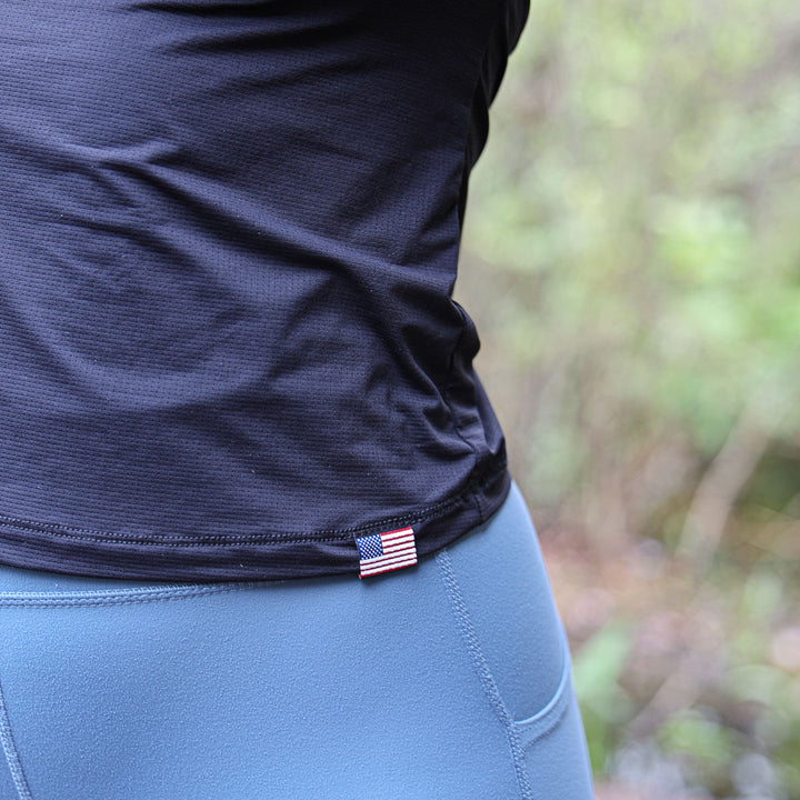 A person in a ToughMesh black shirt with long sleeves and featherweight fabric, paired with blue pants featuring a small US flag tag, stands outdoors.