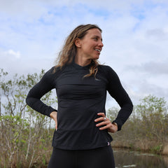 Smiling woman wearing a black long-sleeve athletic top standing outdoors near a wooded wetland under a cloudy sky