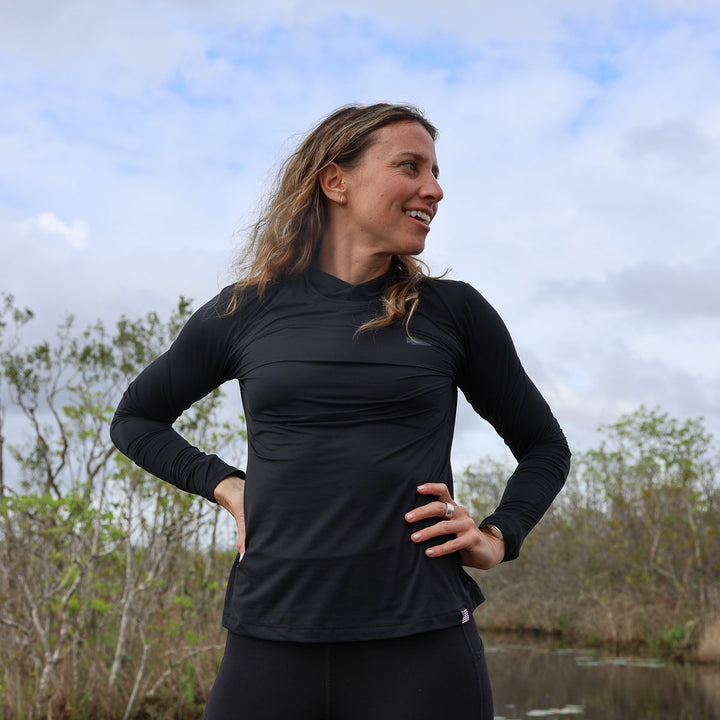 A person confidently stands outdoors in a Women’s Long Sleeve Mock Neck - ToughMesh shirt, shielded by its UV protection, against a backdrop of trees and a cloudy sky.