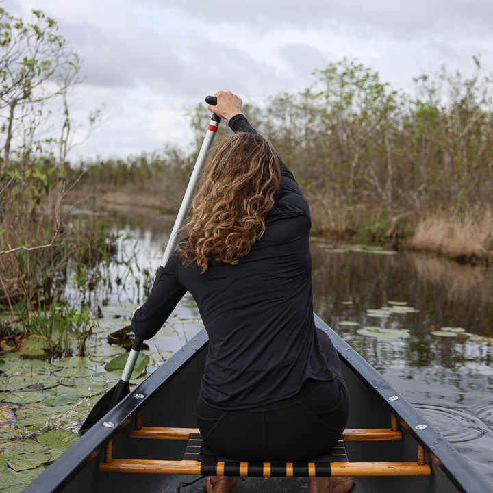 A person paddles through a narrow waterway, surrounded by trees and lily pads, wearing the Women’s Long Sleeve Mock Neck - ToughMesh made from featherweight fabric with UV protection for comfort as they glide smoothly through nature's serene corridor.