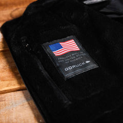 Close-up of a black GORUCK brand "Vest of Power - ToughDry® + Fleece," featuring an American flag label and the text "Proudly Built in the United States of America." This environmentally conscious design is displayed on a wooden surface, representing both sustainability and style.