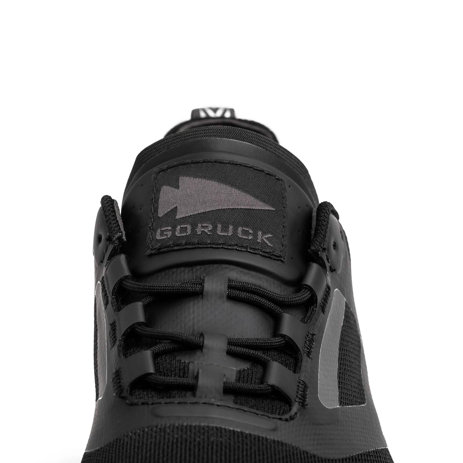 Close-up of a black athletic shoe from GORUCK, featuring the Mackall - Black + Forged Iron design. This sleek trail shoe has black laces and a prominent arrow logo, embodying the durability required for Special Forces training.
