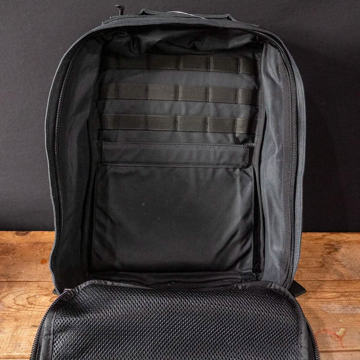 Displayed on a wooden surface with a plain black background, the GR1 Heritage backpack by GORUCK features an open design in black waxed canvas, offering a roomy interior complete with a laptop compartment, multiple straps, and a mesh pocket. It is distinguished by its front panel made of alligator leather.