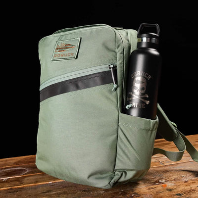 The GORUCK KR1 green kids' rucksack, featuring padded straps and a front zipper pocket with a top patch, holds a black water bottle adorned with a skull and text in its side pocket. This backpack, ideal for school supplies, is placed on a wooden surface against a black backdrop.