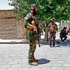 A soldier in camouflage, sporting a beard and helmet, stands firmly with a rifle on a paved area. His feet are clad in GORUCK's MACV-1 Hi-Speed - High Top boots. Behind him, two fellow soldiers rest under a tree by a brick wall, enjoying the sunlit setting.