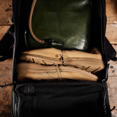 A backpack on a wooden surface is open to reveal its contents, with neatly placed GORUCK MACV-1 Hi-Speed - Mid Top boots inside, soles facing outward, alongside a green hydration bladder.
