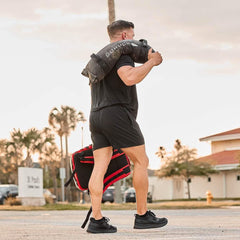 Athletic man carrying a black GORUCK weighted sandbag on shoulder outdoors, wearing black workout gear