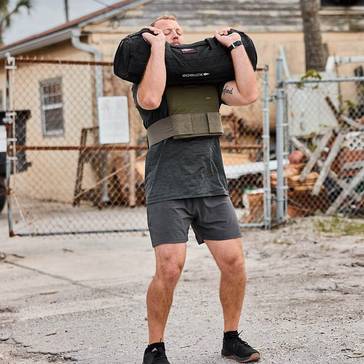 A man is outdoors lifting a heavy sandbag while wearing the durable GORUCK Men’s Training Shorts, made from ToughStretch fabric. His outfit also features a black shirt, green vest, and black shoes, set against a backdrop of a chain-link fence, a house, and various items scattered on the dirt surface.