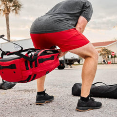 Man in gray shirt and red shorts lifting a red weighted rucksack outdoors on asphalt with palm tree and buildings in background