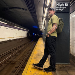 A man wearing the Mackall 45 Black + Gum stands on a subway platform near a “High St Brooklyn Bridge” sign, with a backpack slung over his shoulder.