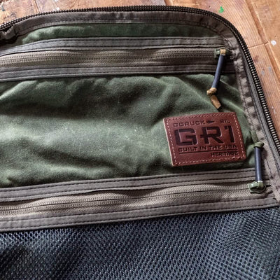 Close-up of a green GORUCK backpack highlighting an alligator leather front panel labeled GR1 Heritage - Built in the USA. This retro canvas rucksack includes zippers and mesh pockets on a wooden surface, embodying timeless style and durability.